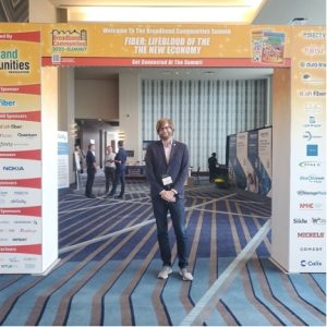 NCC’s Deputy Director, Brian Donoghue, standing under a conference sign reading Welcome to the Broadband Communities Summit; Fiber: The Lifeblood of the New Economy; Get Connected At the Summit.