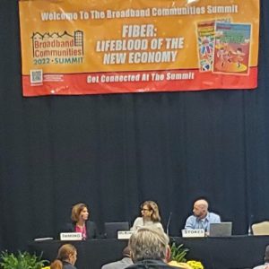 Angela Imming, Director of Technology and Innovation, City of Highland, Illinois; Kim McKinley, Chief Marketing Officer, UTOPIA Fiber; and Sean Stokes, Partner, Keller and Heckman LLP, sitting on a panel under a banner reading Welcome to the Broadband Communities Summit; Fiber: The Lifeblood of the New Economy; Get Connected At the Summit.